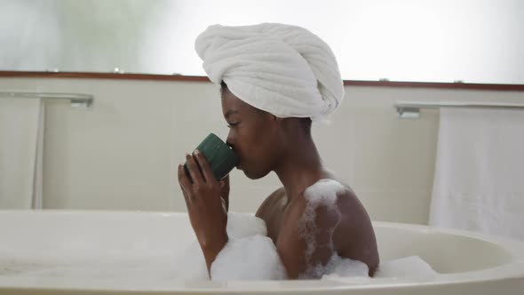 Profile of african american attractive woman relaxing in foam bath and drinking coffee in bathroom alt