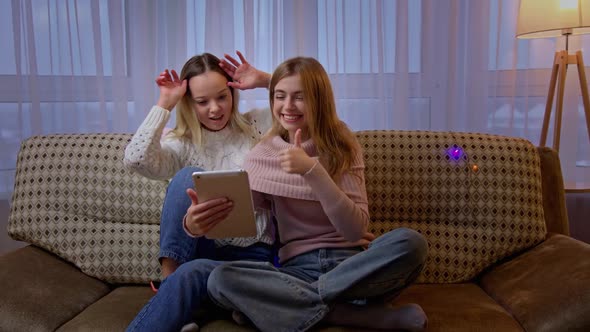 Pretty Young Caucasian Women Sitting on Couch in Living Room and Talking Via Webcam on Tablet Device alt