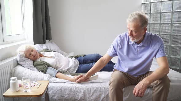 Elderly Woman Is Lying on a Bed and Is Ill and Her Husband Is Sitting Next To Her and Supporting His alt