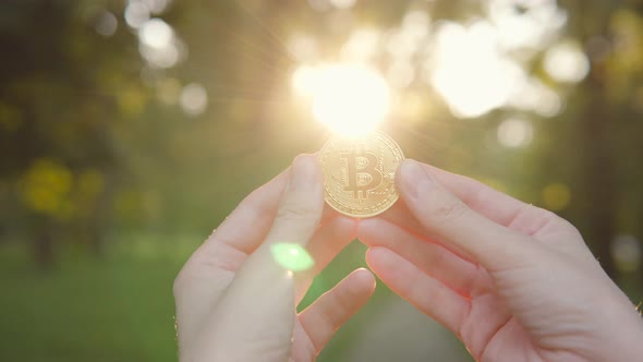 Man Shows a Cryptocurrency Symbol Bitcoin Coin on the Background of a Green Blurred Trees Outdoors