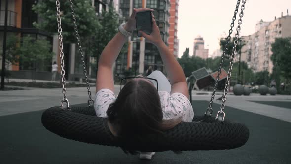 Girl Writes Message on Phonerolls While Lying on Swing