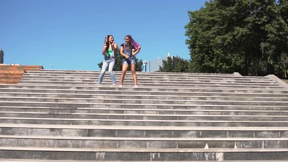 Two Girls Girlfriends are Happy and Jumping Standing on the Steps on the Street alt