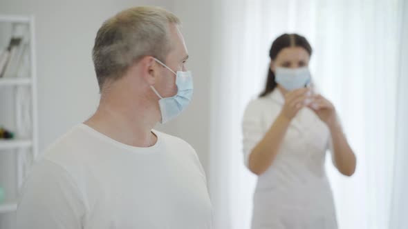 Portrait of Frightened Mid-adult Man in Face Mask Looking Back at Doctor Holding Syringe and Turning alt