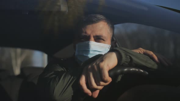 A Sad Man in a Protective Medical Mask Leans on the Steering Wheel in the Car Interior alt