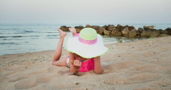 Teen Girl in Beach Hat and Swimsuit is Relaxing Lying on Seashore on Vacation