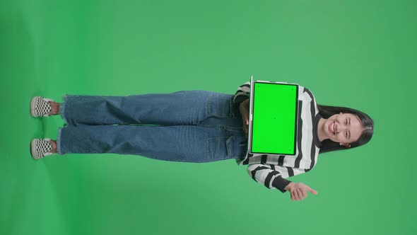 Asian Woman Showing Thumbs Up Gesture While Holding Green Screen Computer In The Green Screen Studio alt