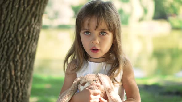 Portrait of Charming Caucasian Girl with Toy Talking Standing in Sunny Summer Park alt