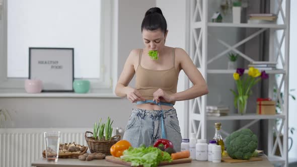 Slim Fit Caucasian Woman Measuring Waist Chewing Salad Leaf Standing at Home Indoors alt
