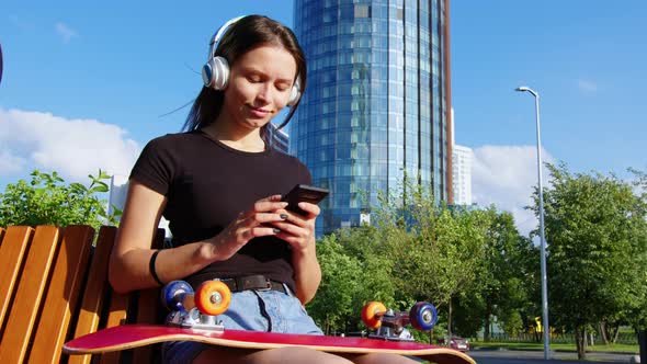 A Woman with a Skateboard Uses a Mobile Phone and Listens to Music alt