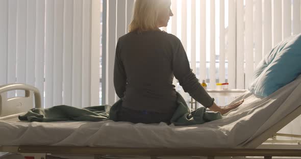 Back View of Senior Female Patient Sitting on Bed in Hospital Ward and Looking Away alt