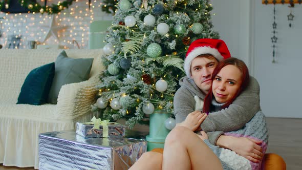 Man and Woman Hugging Tenderly Sitting on Floor Near Christmas Tree in Living Room alt