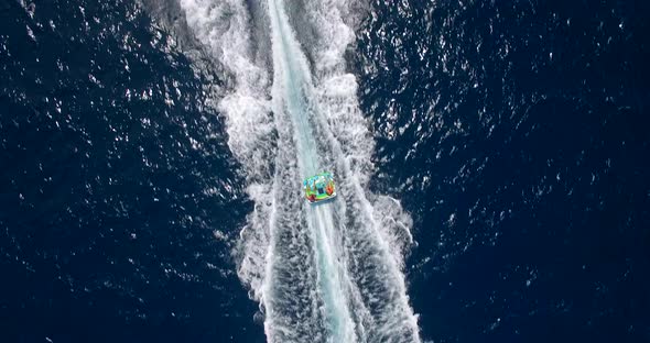 Aerial drone view of man and woman on an inflatable tube towing behind a boat to a tropical island. alt