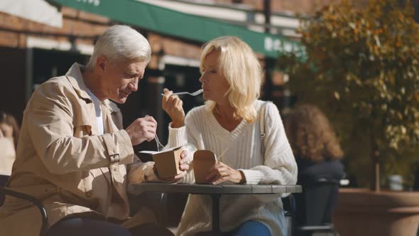 Aged Couple Sitting at Small Outdoor Cafe Table and Enjoying Takeaway Lunch alt