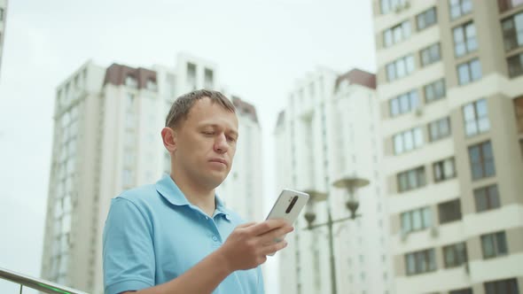 Citizen Surfs in His Mobile Phone Writes a Text Message Stands Background of City Buildings alt