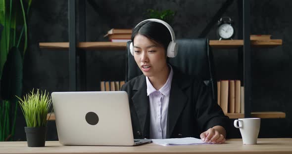Asian Woman in Headphones Using Laptop for Distance Learning and Writes Notes While Sitting at Home alt