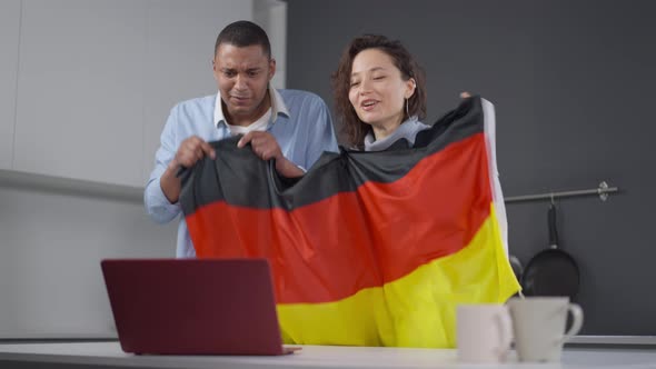 Joyful Interracial Couple with German Flag Cheering for Soccer Team Watching Online Match on Laptop alt