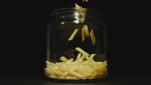 SLOW MOTION: Uncooked Penne Rigate Fall Into Empty Glass Jar On Black Background - Front View alt