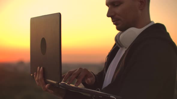 Standing at Sunset on the Roof with a Laptop and Beer in Headphones Work Remotely and Contemplate alt