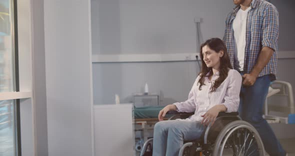 Young Man Pushing Wheelchair with Wife in Hospital Ward alt
