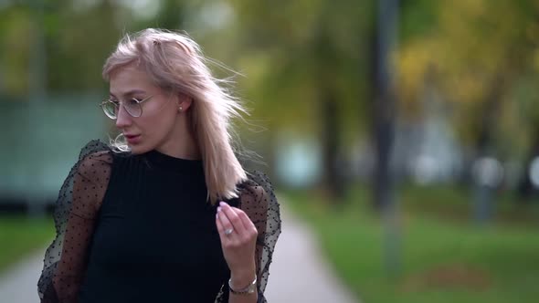A Young Blonde Woman with Glasses Poses Walking Around the City on an Autumn Day in a Park. alt