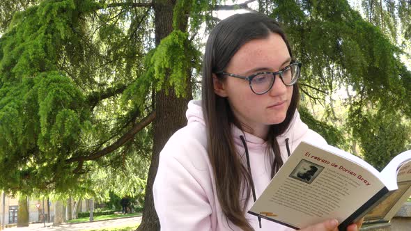 Student girl reading a novel next to a tree on a park, outdoors video. alt