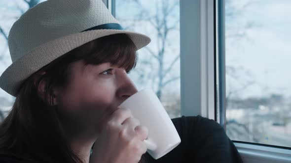 Brunette Woman in Hat Looks Out the Window and Drinks Hot Coffee or Tea  Cinematic Shot