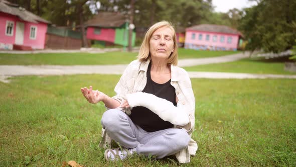 Senior Elderly Woman Doing Yoga Meditation with Injury Broken Arm in Cast Outside in Summer