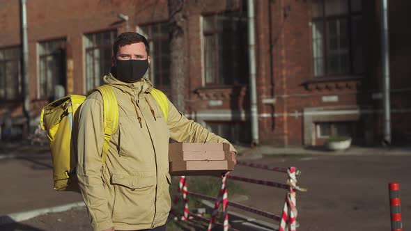 Delivery man in black mask with yellow backpack and pizza boxes takes off mask