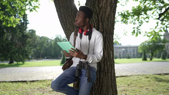 Young Focused African American Student Leaning on Tree Trunk Surfing Internet on Tablet alt