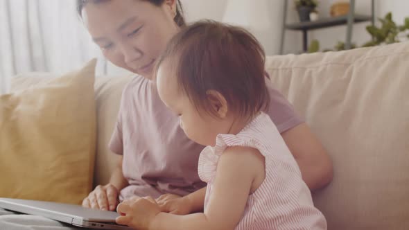 Asian Mom Using Laptop while Sitting with Baby Daughter at Home alt