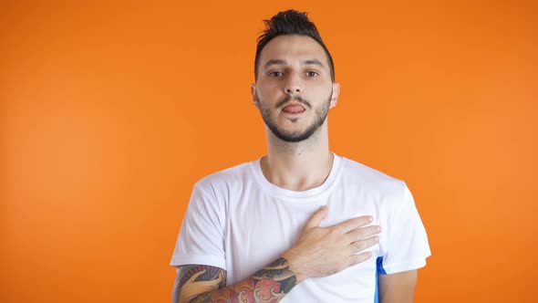 Young Man Football Soccer Fan Singing Hymn with Right Hand on His Heart. Isolated on Orange alt