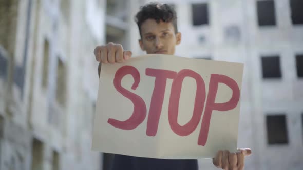 Rack Focus Goes To STOP Banner Raised By Serious Young Man in Slums. Portrait of Concentrated Mixed alt