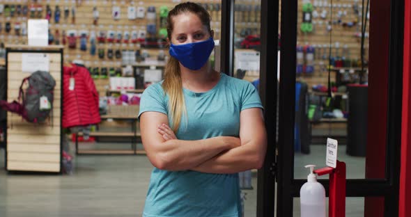 Caucasian female shopkeeper wearing face mask in the doorway of sports shop with arms crossed alt