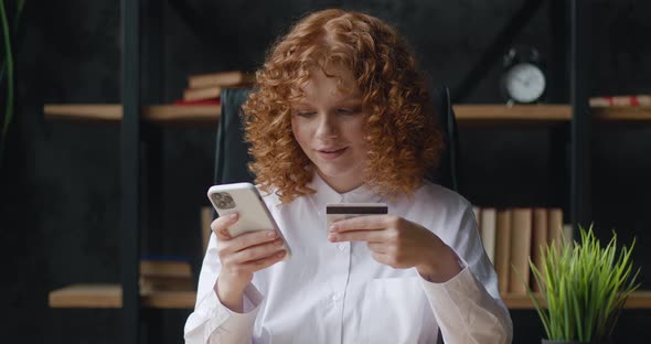 Pretty Redhaired Woman is Making Online Payment Holding Bank Credit Card Using Smartphone at Office alt