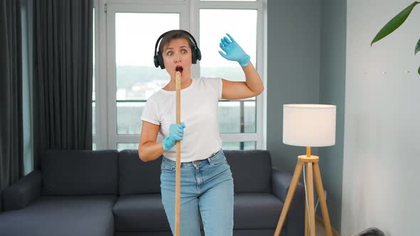 Woman in Headphones Cleans the House and Have Fun Singing with a Broom Like a Star at a Concert alt