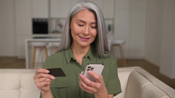 Serene Middleaged Greyhaired Asian Woman Shopping Online Sitting with Credit Card and Mobile Phone