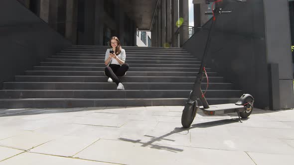 Young Girl Student Sitting on Soda Holding a Mobile Phone. Nearby Is a Modern Transport Electric alt