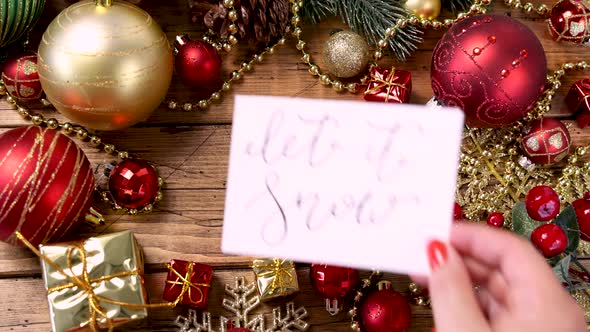 Woman's hand putting a Christmas card with the text LET IT SNOW on a wooden table alt