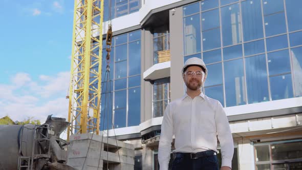 An Engineer in a White Shirt and Helmet Stands Against the Backdrop of a Modern Glass Building alt