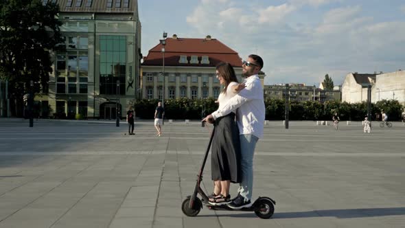 Young Man Rides His Girlfriend on an Electric Scooter Around the City Square