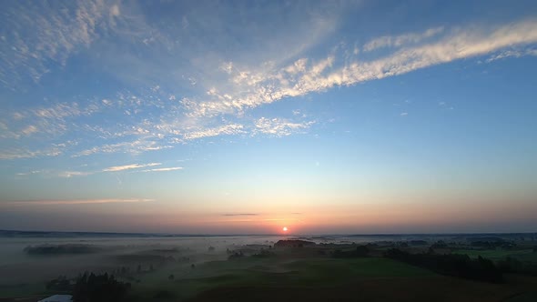 A slow aerial shot of a sunrsie over the field and village in the fog alt