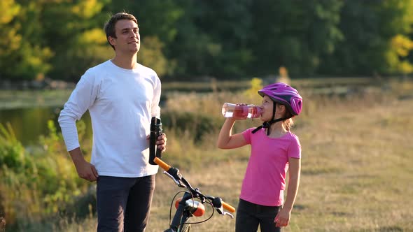 Dad and a little girl in a Bicycle helmet drink clean water near the bike on the river Bank. 