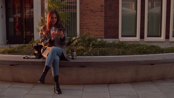 Young attractive business woman sitting outdoor on the bench and using smartphone. alt
