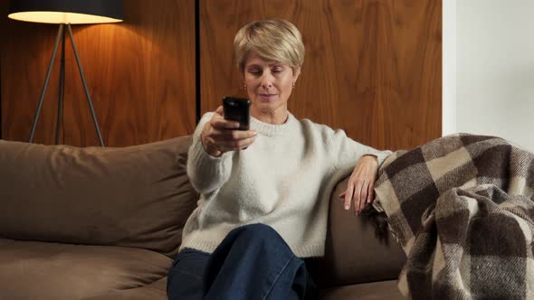 Smiling Middleaged Woman Sitting on the Sofa and Turning on the TV with a Remote Control alt