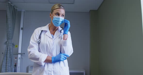 Thoughtful caucasian female doctor face mask and surgical gloves standing at hospital alt