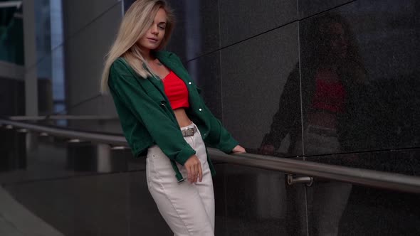 a Blonde in a Red Top and Green Shirt Poses Near a Dark Wall. She's Standing Sideways, Holding on To alt