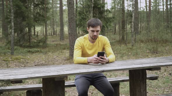 Man with Stubble Sits on Wooden Bench at Table in Forest Looks at Smartphone alt