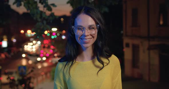 Woman in Glasses and Casual Clothes which Poses on Camera on Lights Night City background alt