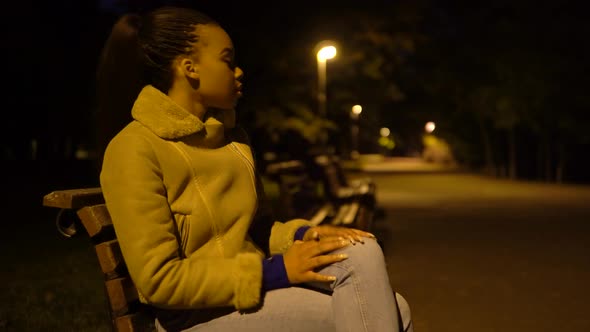 A Young Black Woman is Frustrated As She Sits on a Bench in a City Park at Night  Side View alt