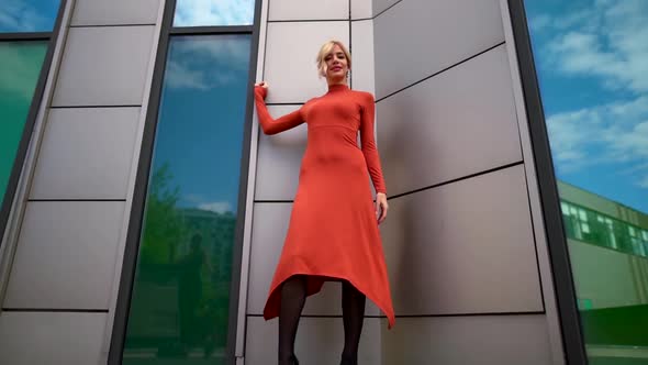 a Blonde in a Red Dress Poses Near Large Green Windows. the Camera Moves From Bottom To Top alt